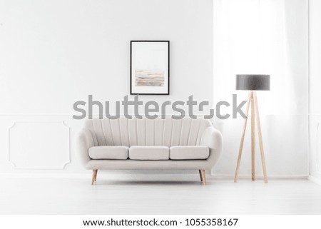 Elegant, beige sofa in an empty white interior with art above the sofa and a wooden tripod lamp by a window