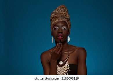 Elegant Black Girl Painting Lips With Lipstick And Looking At Camera. Beautiful Young Slim Woman Wear Traditional African Outfit And Accessories. Isolated On Blue Background. Studio Shoot. Copy