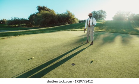 An Elegant Black Guy In A Dapper Outfit With Trousers With Suspenders, Tie, And A Straw Hat, Is Playing Golf On The Field In The Sunset With The Sun Behind Him, Casting A Long Shadow On The Lawn