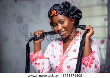 Elegant black lady looking at camera wearing a satin bonnet and floral robe, smiling in interior. Black female health and wellness indoors, skin perfection and care lifestyle.