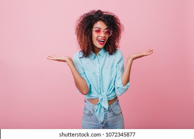 Elegant Black Woman In Casual Attire Expressing Surprised Emotions In Pink Studio. Stunning Short-haired  Girl Posing In Blue Shirt.