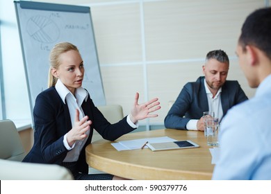 Elegant blond businesswoman gesturing actively while briefing her colleagues at large meeting table in office