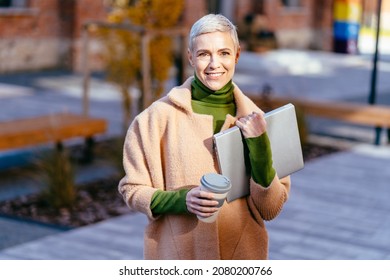 Elegant Blond Short Hair 40-45 Years Old Woman Against Brick Wall Outdoor With Silver Laptop And Takeaway Reusable Coffee Cup In Hands. Remote Working Concept With Flexible Lifestyle.