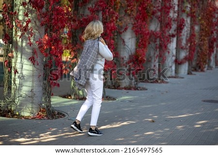 Elegant blonde 35-40 years old in white trousers and jacket walks on foot through autumn city decorated with bright red foliage of overgrown ivy. Walking in fresh air. Urban landscape.
