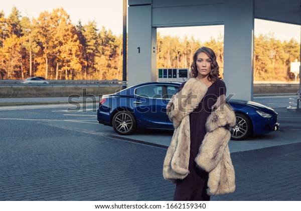 The elegant blonde beautiful woman posing near\
luxury vehicle. Beautiful young woman with blue luxury car. Sexy\
female enjoying trip on luxury modern car. Fashionable lifestyle\
concept.