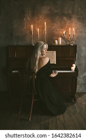 Elegant blonde girl 20-24 year old wearing black vintage style dress playing old piano with burning candles in room closeup.  