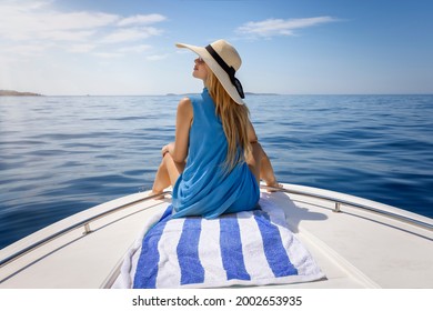 A elegant, blonde woman with a hat sits on a boat and enjoys the calm sea during her summer holidays