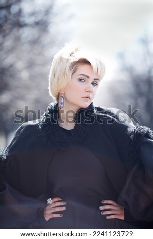 An elegant blonde woman with a short haircut in a black coat on the alley of the park.