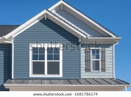 Elegant blue horizontal vinyl siding, shingle and vertical siding in a double gable roof with white decorative corbels, double sash window with matching shutter above a metal roof on a luxury home