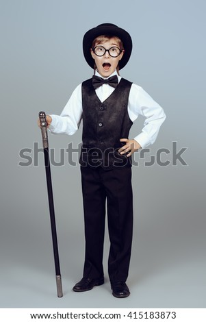 Elegant boy in a suit, bowler hat and glasses posing at studio with a walking stick. Children fashion.