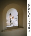 Elegant Bride in White Dress Framed by Arched Window, Holding a Hand Fan, Sitting Gracefully in a Sunlit Room with Minimalist Décor.