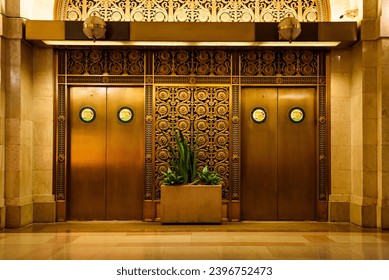 Elegant bronze elevator doors with intricate metalwork and art deco patterns, flanked by marble walls and lush greenery in a planter. - Powered by Shutterstock