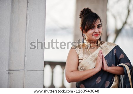 Elegant brunette south asian indian girl in saree standing under arch and show namaste hands sign.
