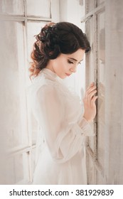 Elegant brunette woman with collected hair in a retro hairstyle stands at the window and is sad. White vintage silk dress. Fine art picture. The mysterious princess in the castle is waiting for prince