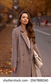 Elegant brunette woman with long wavy hair wearing grey wool coat, holding white handbag and walking at city street on autumn day. Portrait against yellow leaves