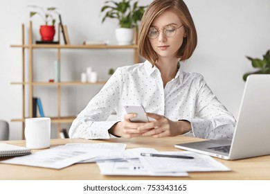 Elegant buisnesswoman dressed formally, surrouded with papers and documents, recieves business message on smart phone, types answer, uses modern electronic gadgets for work and communication