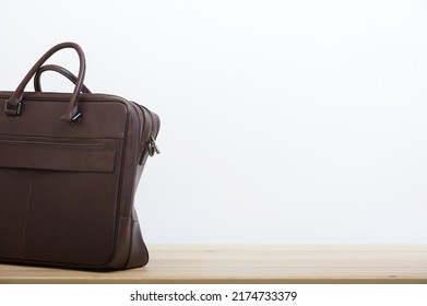 An Elegant Business Briefcase Made Of Old Craft Dark Brown Leather Stands Next To The White Wall As A Background On The Smooth Surface Of Light Wooden Table. No People Studio Photography.