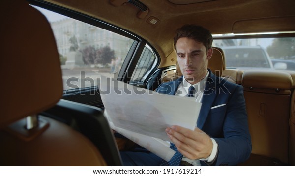 Elegant business man reading documents in\
business car. Focused male professional reviewing statistics in\
paperwork in modern car. Handsome businessman working with papers\
in automobile.