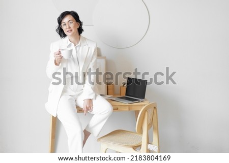 Elegant business woman with black hair, middle-aged in a white business suit. Holds a cup of coffee in his hand. Cozy office with wooden furniture. Copy space.