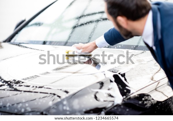 Elegant businessman\
dressed in the suit washing his car with yellow sponge and foam on\
a self service car wash
