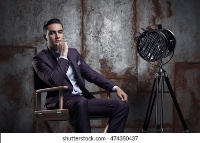 Elegant Businessman In Fashionable Suit Posing In The Photo Studio . Luxury Portrait Of A Man Sitting In The Director's Chair Next To The Model Is A Retro Movie Spotlight