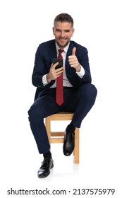 Elegant Businessman In Suit With Telephone Scrolling On Instagram And Making Thumbs Up Gesture While Sitting On Wooden Chair On White Background