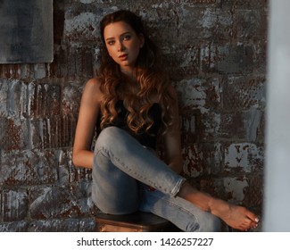 Elegant casual dressed woman sitting at high chair against brick wall. Girl in jeans and black top with long curly hair. Loft interior. Evening sun light. Full length portrait