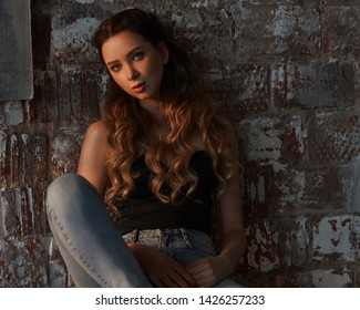 Elegant casual dressed woman sitting at high chair against brick wall. Girl in jeans and black top with long curly hair. Loft interior. Evening sun light