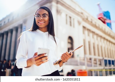 Elegant Cheerful African American Woman In Glasses And White Shirt Walking With Mobile Phone Tablet And Documents In Hand Looking At Camera In Sunny Street In New York City