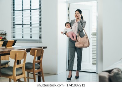 Elegant Chinese Businesswoman Just Arriving Home With Kid Is Answering Her Customer's Call. Happy Asian Career Woman Carrying Infant Is Having Peaceful Conversation With Nanny On Cellphone.