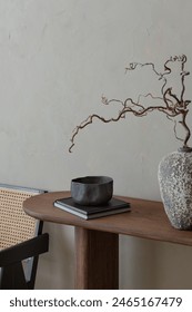 Elegant composition of warm living room interior with wooden console, black bowl, books, stylish vase with branch, rattan armchair, beige wall and personal accessories. Home decor. Template.