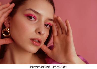 Elegant confident beautiful young woman with bold fuchsia color eyeshadow eyes makeup, flawless, glow skin, wearing trendy earrings, posing on pink background. Close up studio beauty portrait