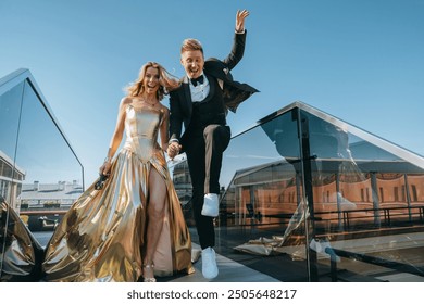 Elegant couple in formal attire, joyfully jumping and holding hands on a rooftop, the woman in a gold gown and the man in a tuxedo, celebrating under a clear blue sky - Powered by Shutterstock