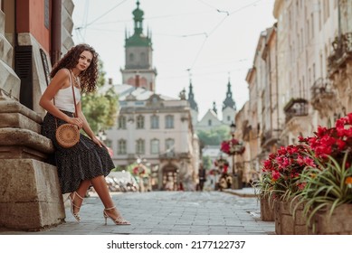 Elegant curly brunette woman wearing trendy summer outfit with round wicker shoulder bag, white top, polka dot midi skirt, strap sandals, posing in street of European city. Copy, empty space for text - Shutterstock ID 2177122737