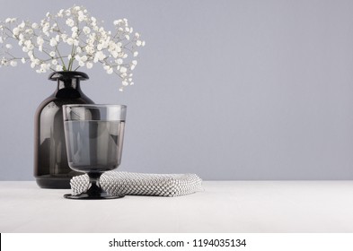Elegant decor dressing table in minimalist style - black vase with flowers, glass, cosmetic accsssories silver bag on grey wall and white wood background.