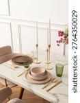 Elegant dining setup with marble table, gold cutlery, pink and green dishware, tall candles, and floral decor in a modern setting.