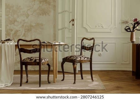 An elegant dining space featuring two antique wooden chairs with patterned upholstery, a simple light wood table, and decorative walls and floor