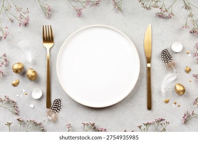 Elegant Easter table composition with empty white plate, golden cutlery, gold eggs, feathers and pink spring flowers on light stone background. Festive flat lay with copy space.