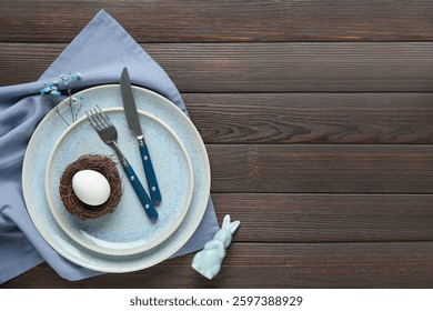 Elegant easter table setting with nest and egg, cutlery, and bunny figurine on dark wooden background