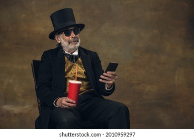 Elegant Elderly Gray-haired Man, Gentleman Or Actor Using Phone Isolated On Dark Vintage Background. Retro Style, Comparison Of Eras Concept. Old Male Model Like Aristoctar.