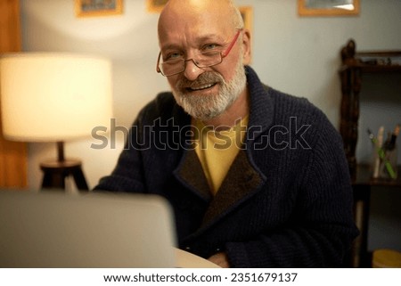Elegant elderly male with grey beard in glasses and stylish cardigan sitting at home in his cabinet in front of laptop, looking at camera with warm smile. Senior male author writing novel on computer