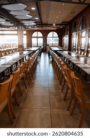 Elegant empty restaurant interior with long wooden tables and rows of matching chairs prepared for a group event or banquet, warm sunlight shining through arched windows; wide-angle vertical shot