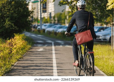 Elegant entrepreneur in smart casual cycling on bicycle track in warm sunny day. Back view of tall male manager with brown leather laptop bag riding bike in sleeping area of city. Concept of activity. - Powered by Shutterstock