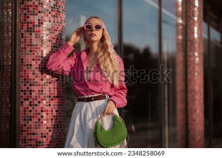 Elegant fashionable blonde woman wearing trendy summer outfit with pink sunglasses, linen shirt, belt, white bermuda shorts, holding small green faux leather bag, posing in street of city. Copy space