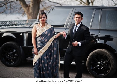 Elegant And Fashionable Indian Friends Couple Of Woman In Saree And Man In Suit Posed Against Rich Black Suv Car.