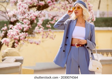 Elegant fashionable woman wearing trendy outfit: blue suit with blazer, classic trousers, white crop top, belt, beret, holding white leather bag, posing in street, near blooming tree. Copy space