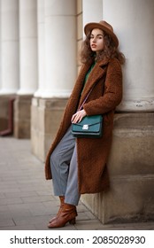 Elegant Fashionable Woman Wearing Trendy Midi Brown Faux Fur Teddy Bear Coat, Hat, Checked Trouthers, Ankle Boots, With Green Leather Shoilder  Bag, Posing In City. Street Style Full-length Portrait