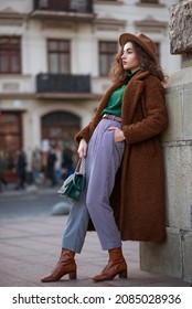 Elegant Fashionable Woman Wearing Trendy Midi Brown Faux Fur Teddy Bear Coat, Hat, Checked Trouthers, Ankle Boots, Holding Green Leather Bag, Posing In City. Street Style Full-length Portrait