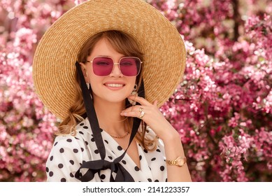 Elegant fashionable woman wearing trendy straw hat, pink square sunglasses, wrist watch, polka dot blouse, posing in street near pink spring blossom trees. Outdoor lifestyle portrait. Copy space  - Shutterstock ID 2141223357