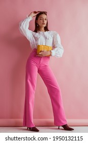 Elegant fashionable woman wearing white vintage blouse with lace collar, pink jeans, holding small padded yellow leather bag, posing on pink background. Full length portrait
				